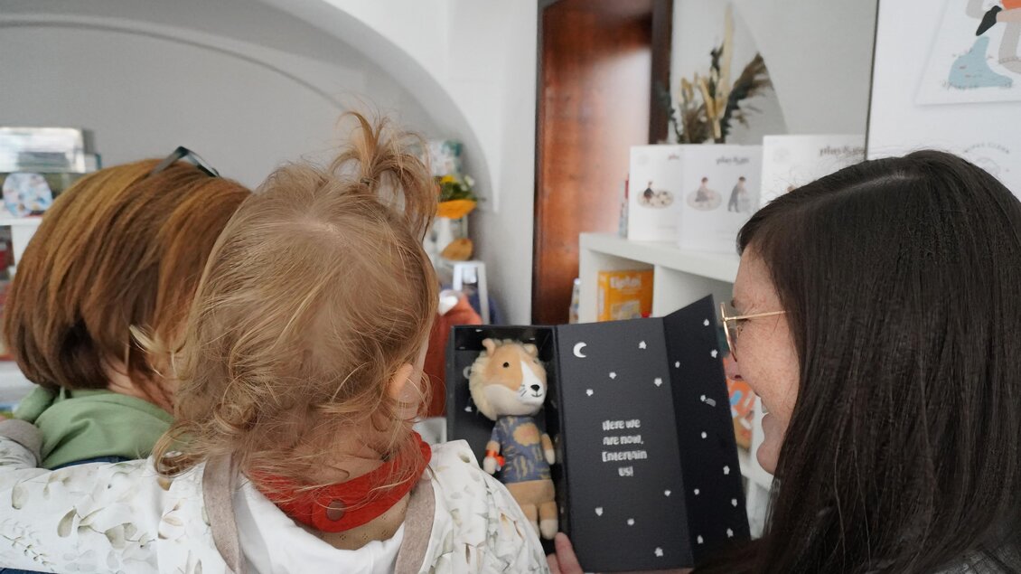 Eine Mutter hält ein Kind, während sie ein Kinderbuch mit einem Spielzeugbear vorlesen. Im Hintergrund sind farbenfrohe Produkte und eine gemütliche Ladenatmosphäre zu sehen. | © Denise Siedler