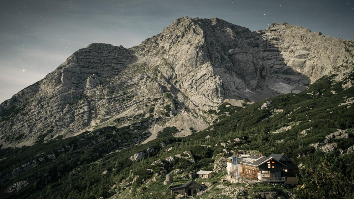 Eine einsame Berghütte liegt in einer grünen Landschaft unterhalb einer beeindruckenden Bergkette. Der Himmel ist klar und die Umgebung ist geprägt von Felsen und Wäldern. | © Stefan Leitner