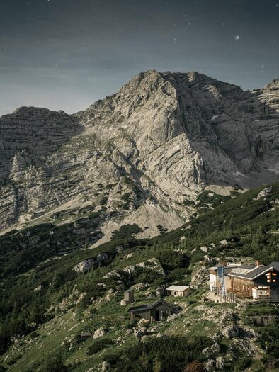 A lonely mountain cabin is situated in a green landscape beneath an impressive mountain range. The sky is clear, and the surroundings are characterized by rocks and forests. | © Stefan Leitner