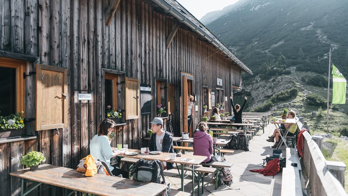 Eine Holzterrasse mit Tischen und Stühlen, wo Menschen entspannen und essen. Im Hintergrund sind Berge und ein klarer Himmel zu sehen. | © Stefan Leitner