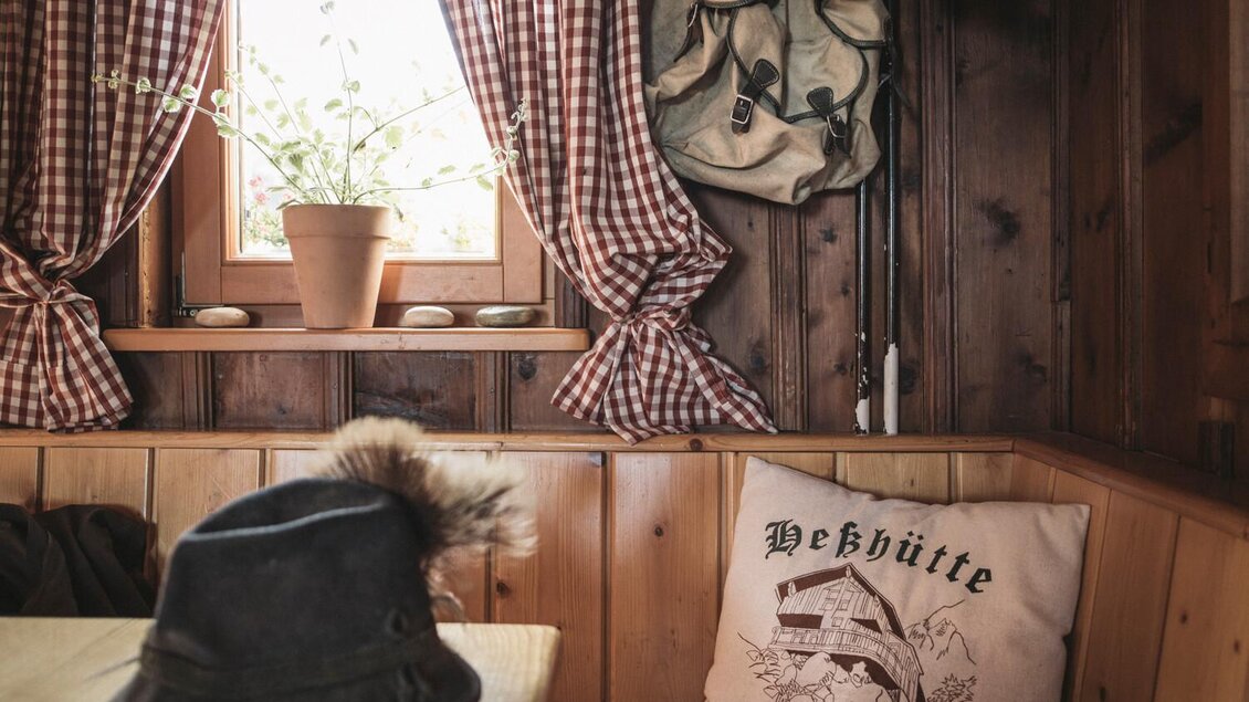 Ein gemütlicher Innenraum mit Holzwänden und karierten Vorhängen. Auf dem Tisch liegt ein Hut und ein Kissen mit dem Aufdruck "Hebhütte Gesäuse". | © Stefan Leitner