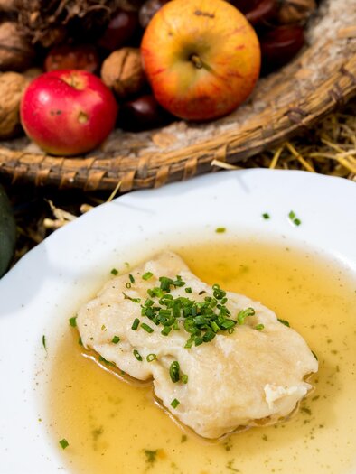 Ein Teller mit einer traditionellen Speise in Brühe, garniert mit frischem Schnittlauch. Im Hintergrund sind Äpfel, Nüsse und Maiskolben in einem Korb zu sehen. | © Region Graz-Harry Schiffer