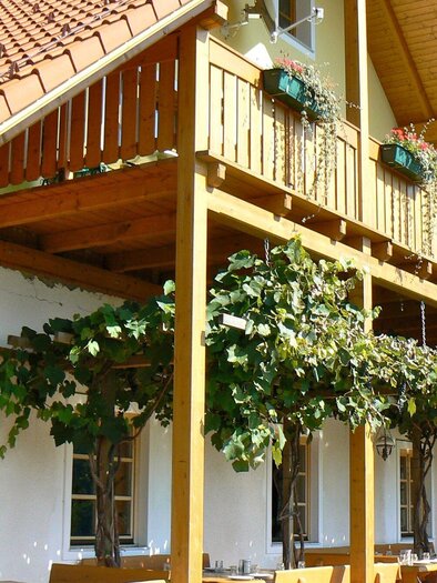 Ein charmantes Haus mit einem Holzbalkon, der mit Pflanzen dekoriert ist. Die umliegende Architektur ist hell und einladend. | © Heurigenschenke Sattler