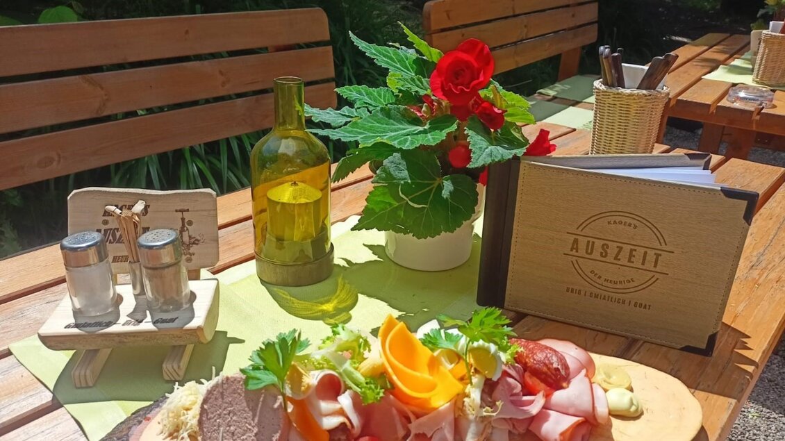 Tisch gedeckt im Außenbereich mit einem Holzbrett, auf dem eine Auswahl an Aufschnitt, Käse und frischem Gemüse serviert wird. Im Hintergrund ein Menübuch mit der Aufschrift „AUSZEIT“ und eine Flasche Öl auf einem Tisch unter Sonnenschein. | © Kager's Auszeit