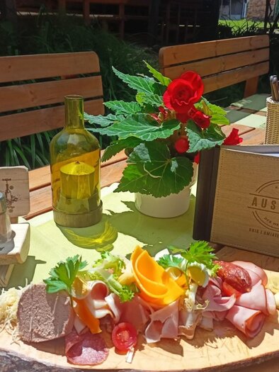 Tisch gedeckt im Außenbereich mit einem Holzbrett, auf dem eine Auswahl an Aufschnitt, Käse und frischem Gemüse serviert wird. Im Hintergrund ein Menübuch mit der Aufschrift „AUSZEIT“ und eine Flasche Öl auf einem Tisch unter Sonnenschein. | © Kager's Auszeit