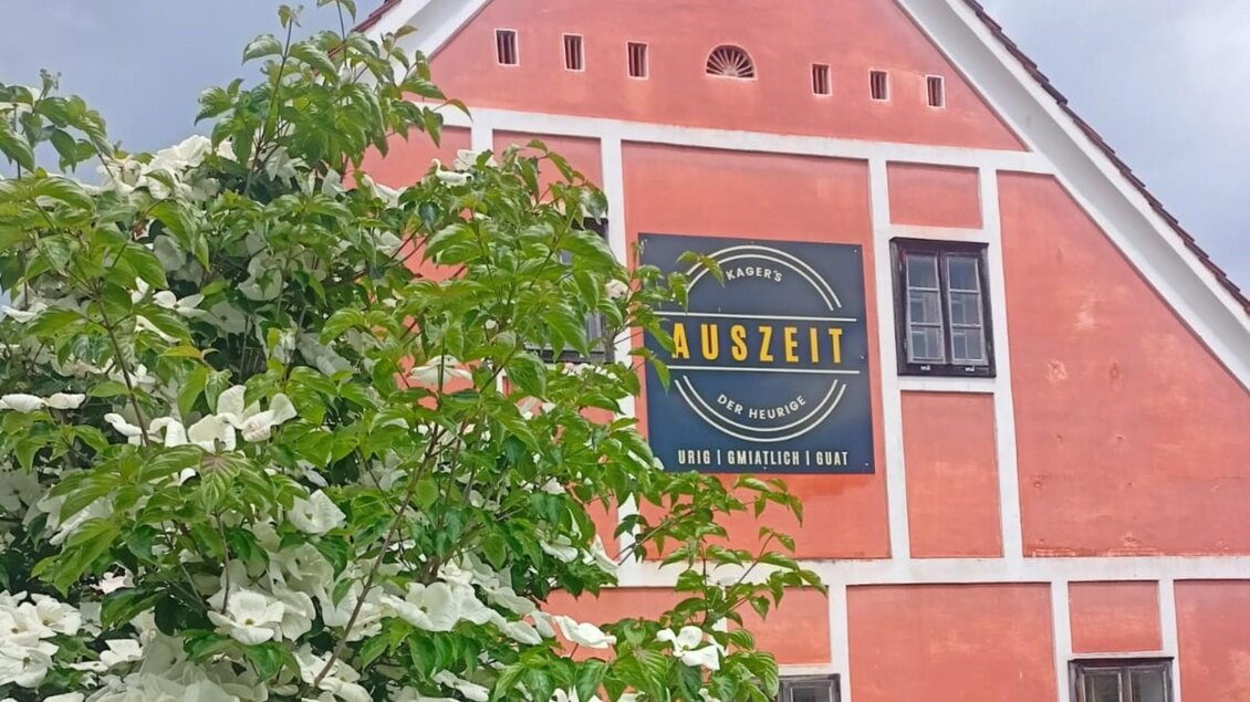 Fassade eines Hauses in kräftigem Orange mit einem großen Schild, auf dem „Kager’s AUSZEIT – DER HEURIGE“ steht. Vor dem Gebäude blüht ein Strauch mit weißen Blüten. | © Kager`s AUSZEIT
