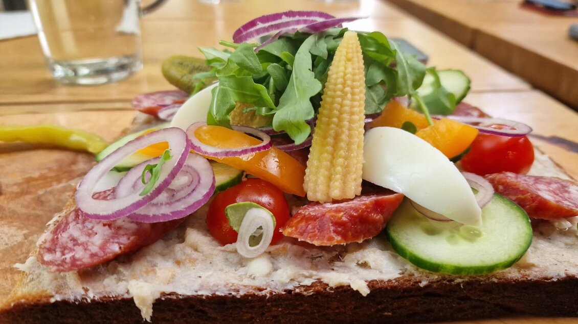 Ein belegtes Brot mit Wurst, frischem Gemüse wie Tomaten, Gurken, Zwiebeln, Paprika, kleinen Maiskolben und einem hartgekochten Ei, garniert mit Rucola. Ein Glas Wasser ist im Hintergrund sichtbar. | © Kager`s AUSZEIT