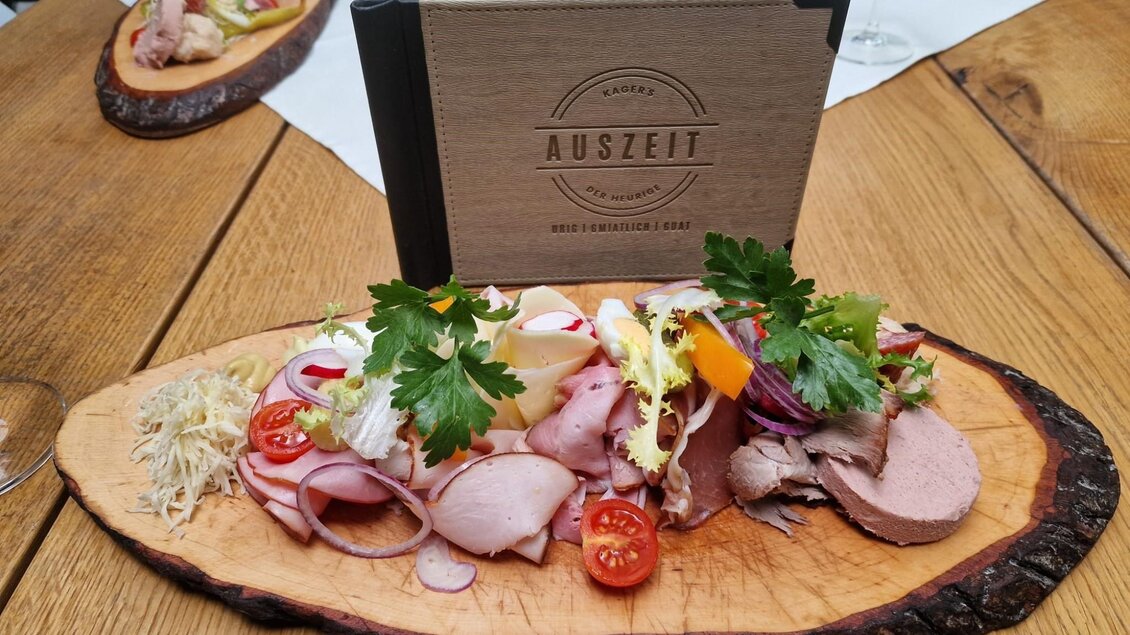 Ein rustikal serviertes Brett mit verschiedenen Wurst- und Schinkenaufschnitten, frischen Tomaten, Zwiebeln, Gemüse und einer Portion Krautsalat. Im Hintergrund ein „AUSZEIT“-Menübuch und ein Glas Wein. | © Kager`s AUSZEIT