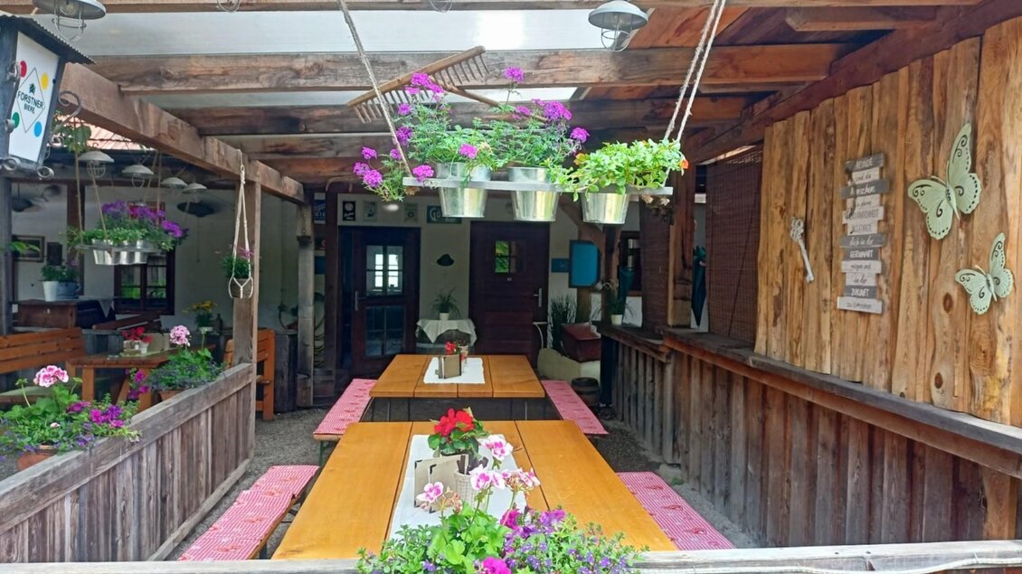 Blick auf eine gemütliche überdachte Terrasse mit Holzverkleidung und Blumenpflanzen in Töpfen. Auf den Tischen stehen Pflanzenarrangements, und es hängen Lampen sowie Blumenkästen von der Decke. An der Wand ist eine Dekoration mit Schmetterlingen und einem Holzbalken mit Sprüchen zu sehen. | © Kager`s AUSZEIT