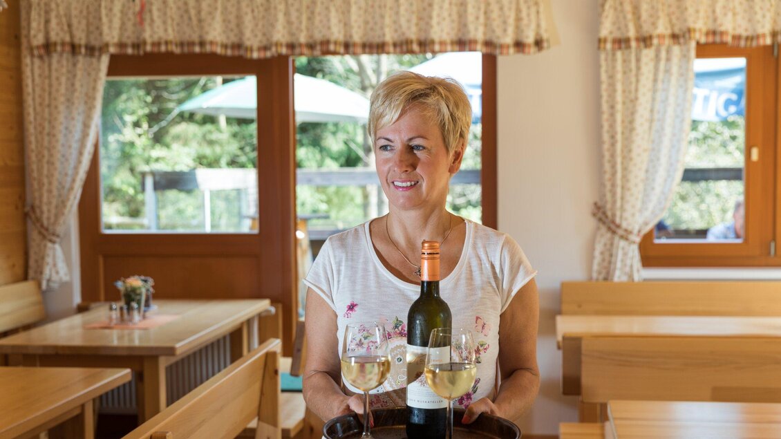 Eine lächelnde Frau trägt ein Tablett mit zwei Gläsern Wein und einer Flasche. Die Umgebung ist ein einladendes Restaurant mit Holzsitze und Fenstern zum Außenbereich. | © TV Region Graz - Harry Schiffer