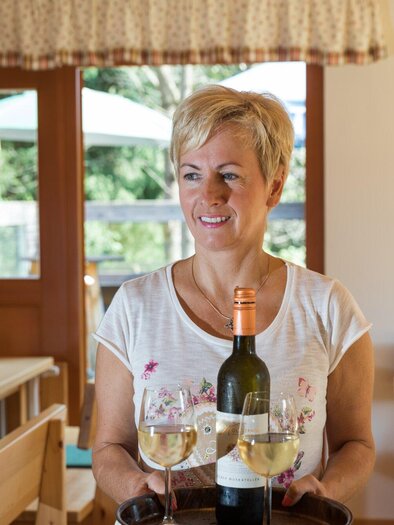 Eine lächelnde Frau trägt ein Tablett mit zwei Gläsern Wein und einer Flasche. Die Umgebung ist ein einladendes Restaurant mit Holzsitze und Fenstern zum Außenbereich. | © TV Region Graz - Harry Schiffer