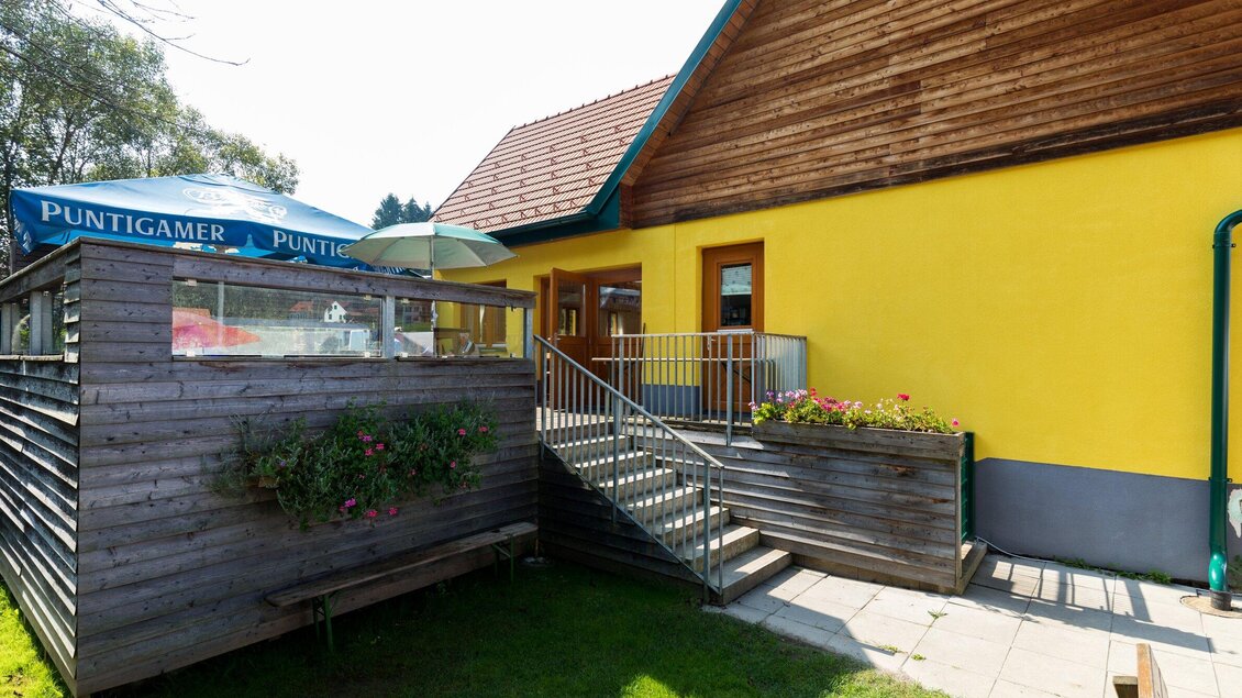 Ein gemütliches Gebäude mit gelber Fassade und Holzverkleidung. Der Außenbereich hat eine Terrasse mit Sonnenschirmen und blühenden Pflanzen. | © TV Region Graz - Harry Schiffer