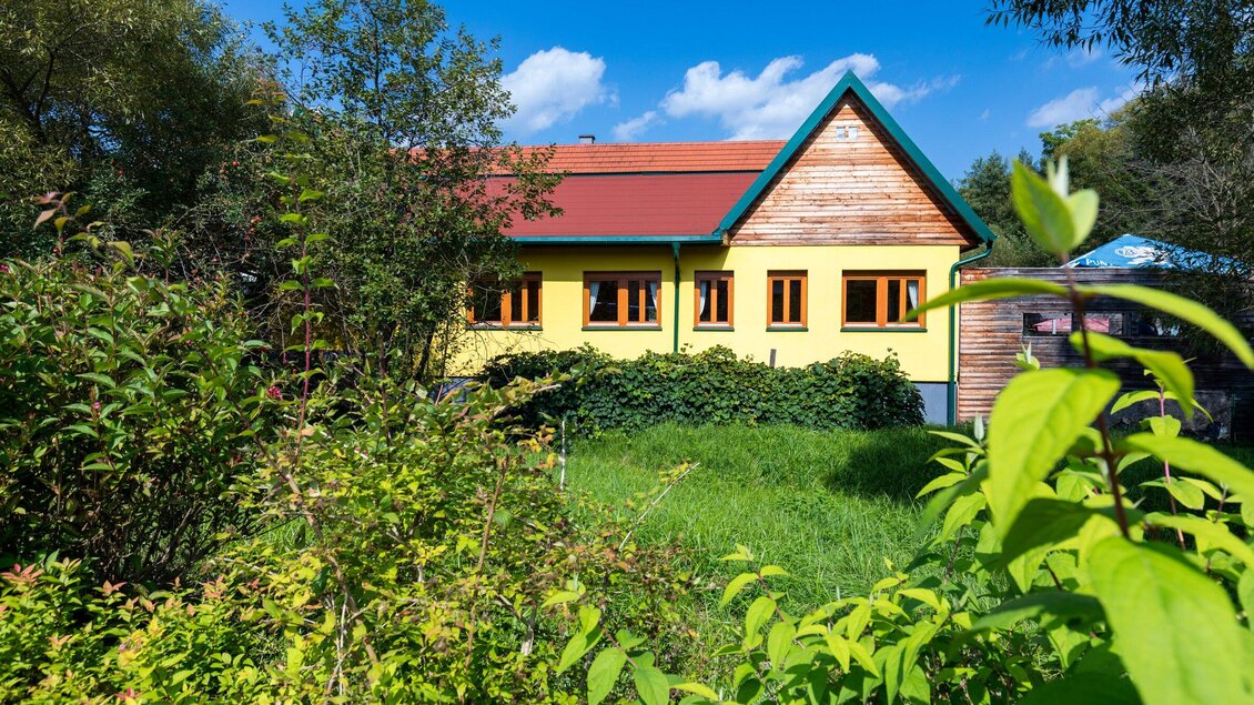 Ein buntes Haus mit einem roten Dach und großen Fenstern. Umgeben von üppigem Grün und strahlend blauem Himmel. | © TV Region Graz - Harry Schiffer