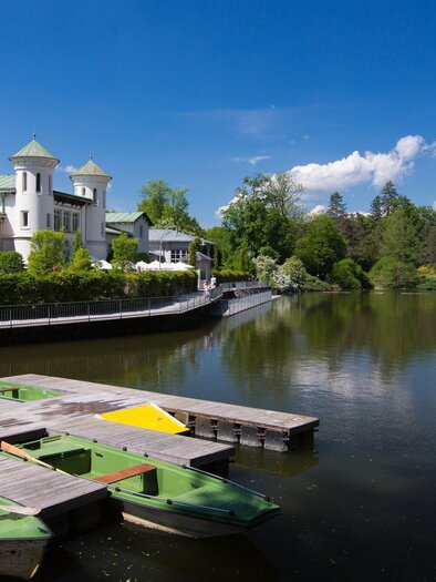 At the dark shimmering Hilmteich in Graz with a boat dock featuring rowing boats, the historical building of Café Purberg is visible in the background on a sunny summer day. | © Graz Tourismus-Harry Schiffer