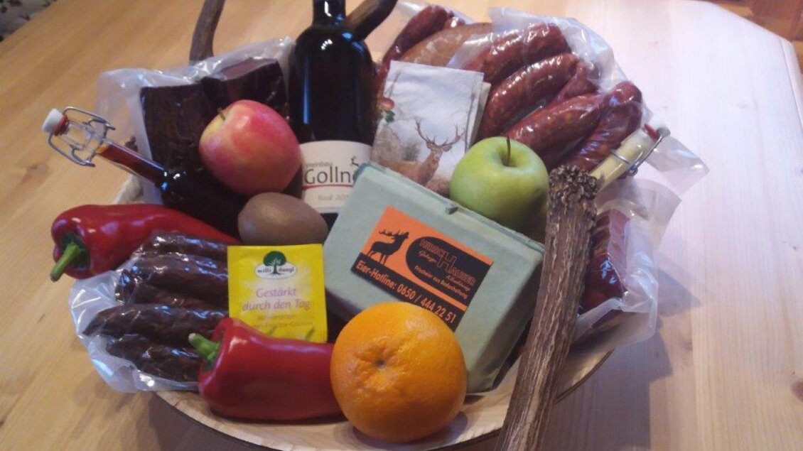 Ein Präsentkorb mit verschiedenen Leckereien wie Wurst, Käse, Obst und einer Flasche Wein. Der Korb ist dekorativ angeordnet und bietet eine Vielfalt an regionalen Spezialitäten. | © Hirschgehege Hauser