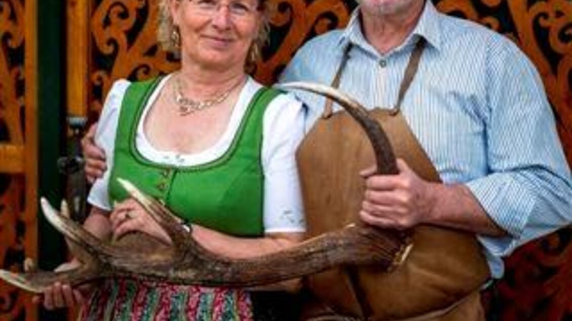 Ein Paar steht vor einer kunstvoll verzierten Wand. Sie tragen traditionelle Kleidung und halten Geweihstücke in den Händen. | © Hirschhorn Diethart