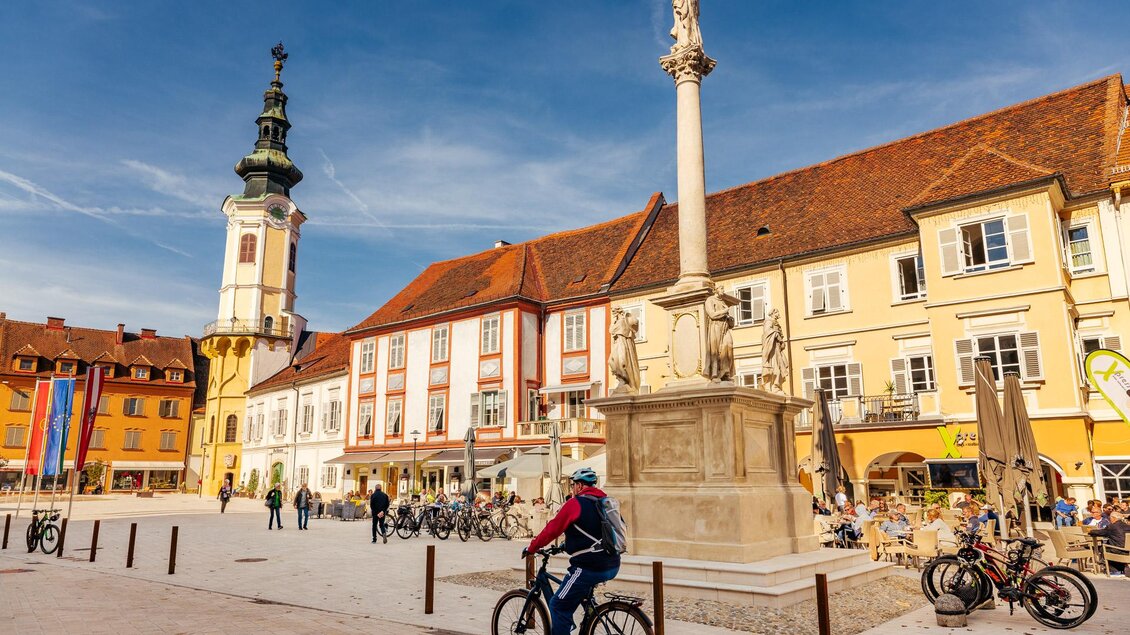 Der belebte Hauptplatz von Bad Radkersburg zeigt bei strahlendem Sonnenschein historische Gebäude, den Rathausturm, die Mariensäule und Radfahrer:innen sowie Gäste in Cafés – ein einladendes Bild steirischer Altstadtkultur. | © Zehnerhaus Bad Radkersburg