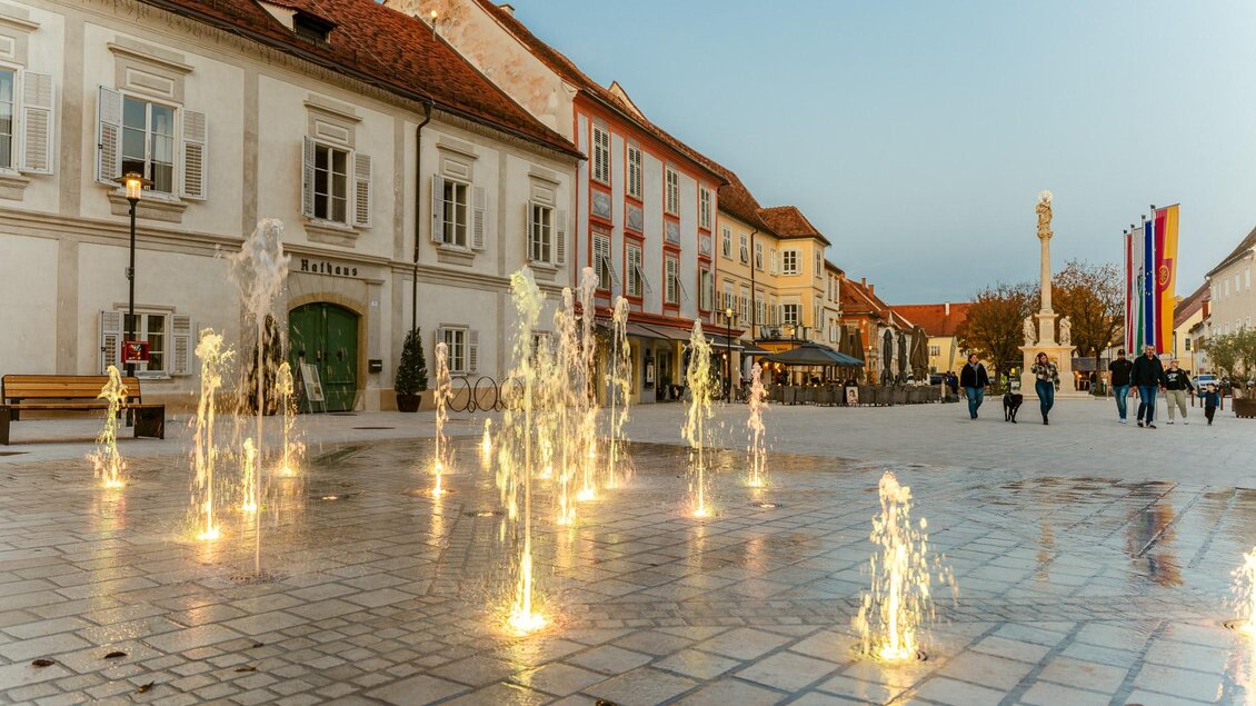 Im sanften Abendlicht sorgen beleuchtete Wasserfontänen auf dem Hauptplatz von Bad Radkersburg vor historischen Fassaden und der Mariensäule für eine stimmungsvolle Atmosphäre, während Menschen gemütlich flanieren. | © Zehnerhaus Bad Radkersburg