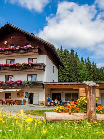 Ein gemütliches Landhaus umgeben von Wiesen und Bäumen. Bunte Blumen schmücken die Fensterbalkone und die sonnige Umgebung.