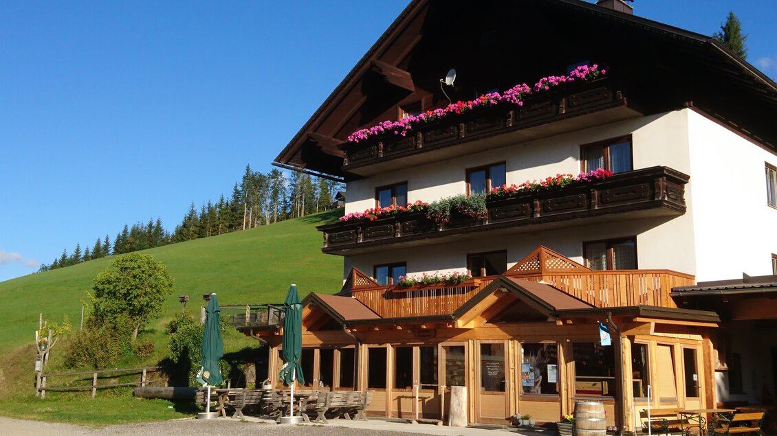 Ein charmantes Landhaus mit bunten Blumen auf dem Balkon. Im Vordergrund befindet sich eine einladende Terrasse mit grünen Sonnenschirmen. | © Schäffer