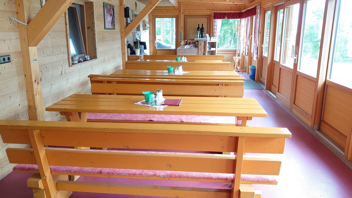 Ein gemütlicher Innenraum mit Holztischen und Bänken. Große Fenster lassen viel Licht herein und bieten einen Blick nach draußen.