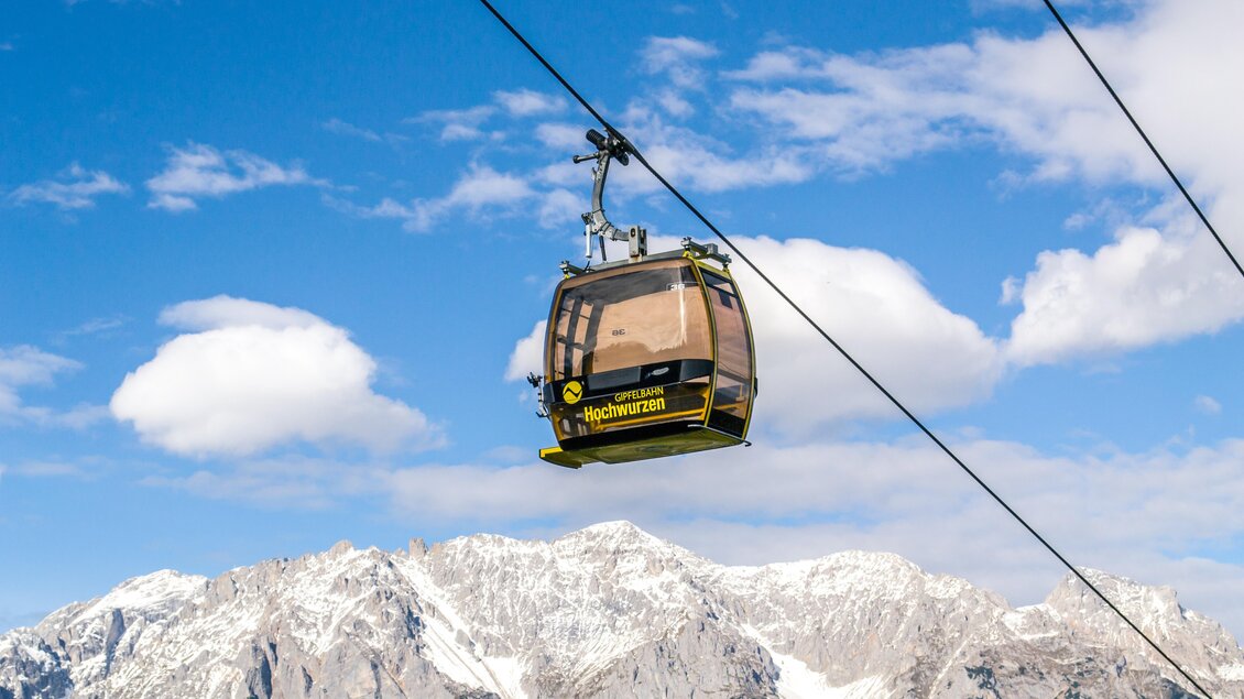 Eine Seilbahn schwebt über schneebedeckte Berge unter einem klaren blauen Himmel. Im Hintergrund sind weiße Wolken zu sehen. | © Planai/Klünsner