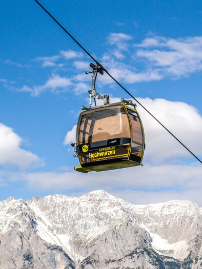 A cable car glides over snow-covered mountains under a clear blue sky. In the background, white clouds can be seen. | © Planai/Klünsner