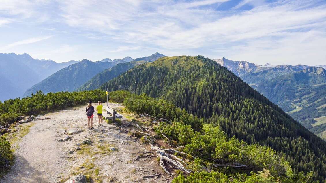 Eine atemberaubende Berglandschaft mit grünen Hügeln und hohen Gipfeln. Zwei Wanderer genießen die Aussicht auf dem Weg. | © Josh Absenger