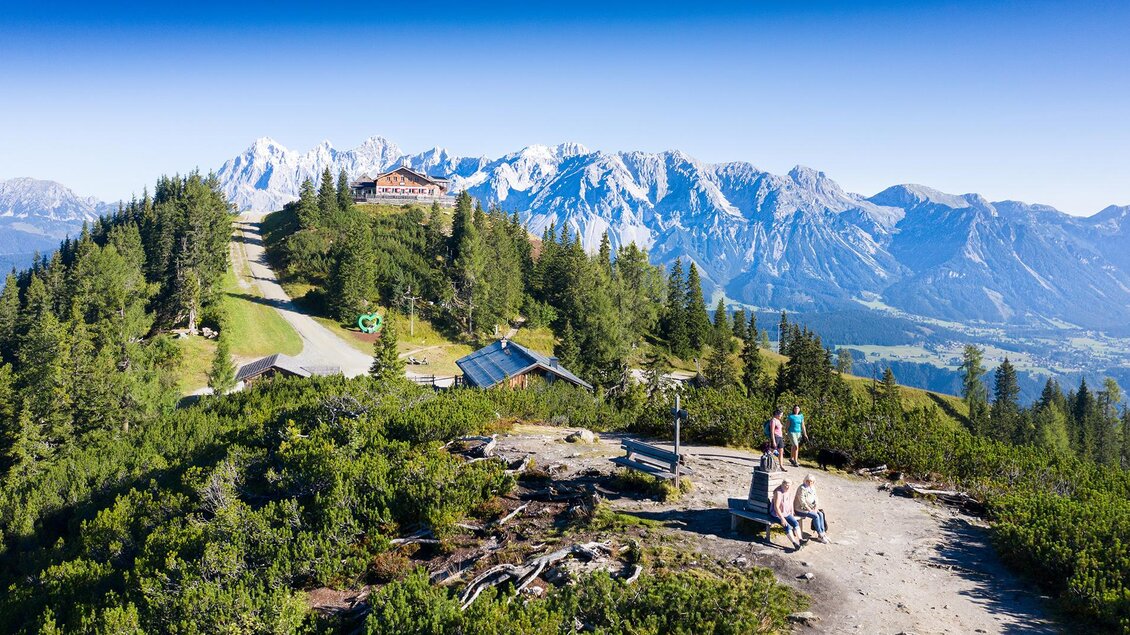 Eine atemberaubende Berglandschaft mit schneebedeckten Gipfeln und grünen Bäumen. Ein Wanderweg und eine Berghütte sind gut sichtbar. | © Josh Absenger