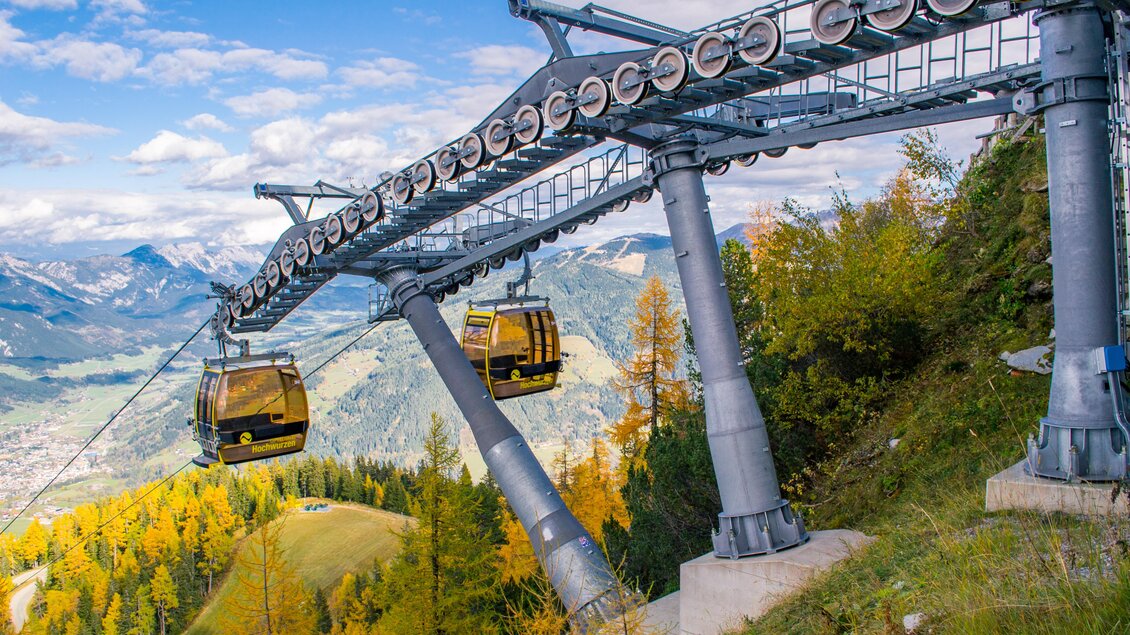 Eine Seilbahn fährt über einen malerischen Berg. Die Landschaft zeigt bunte Bäume und eine beeindruckende Bergkulisse. | © Planai/Klünser