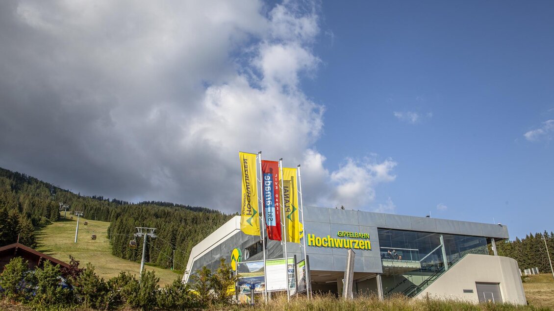 Ein modernes Gebäude mit farbigen Fahnen steht vor einem Bergpanorama. Im Hintergrund sind die Skilifte und die bewaldeten Hänge sichtbar. | © Planai/Kovacsics