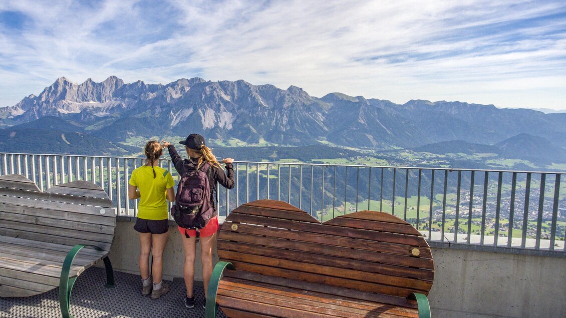 Zwei Personen stehen auf einer Aussichtsplattform und blicken auf die Berge. Im Vordergrund sind Bänke zu sehen, darunter eine herzförmige Bank. | © Johannes Absenger