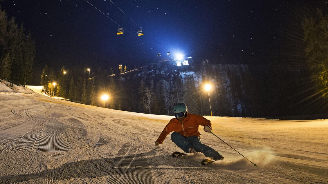 Nacht-Skifahren auf einer beleuchteten Piste mit einem Skifahrer in Bewegung. Die Lifte sind im Hintergrund sichtbar, umgeben von Bäumen und Sternen. | © Gregor Hartl