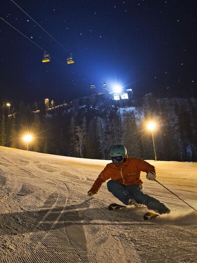 Nacht-Skifahren auf einer beleuchteten Piste mit einem Skifahrer in Bewegung. Die Lifte sind im Hintergrund sichtbar, umgeben von Bäumen und Sternen. | © Gregor Hartl