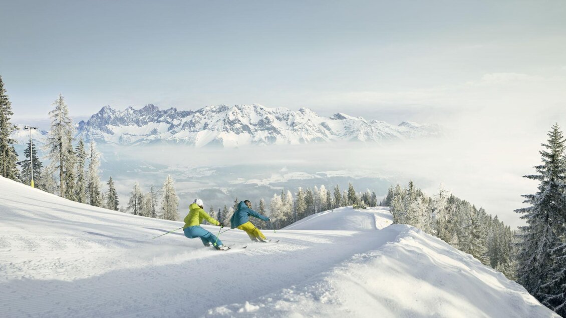 Zwei Skifahrer fahren durch unberührten Snow auf einem Berg. Im Hintergrund sind schneebedeckte Gipfel und eine Nebelschicht zu sehen. | © Peter Burgstaller