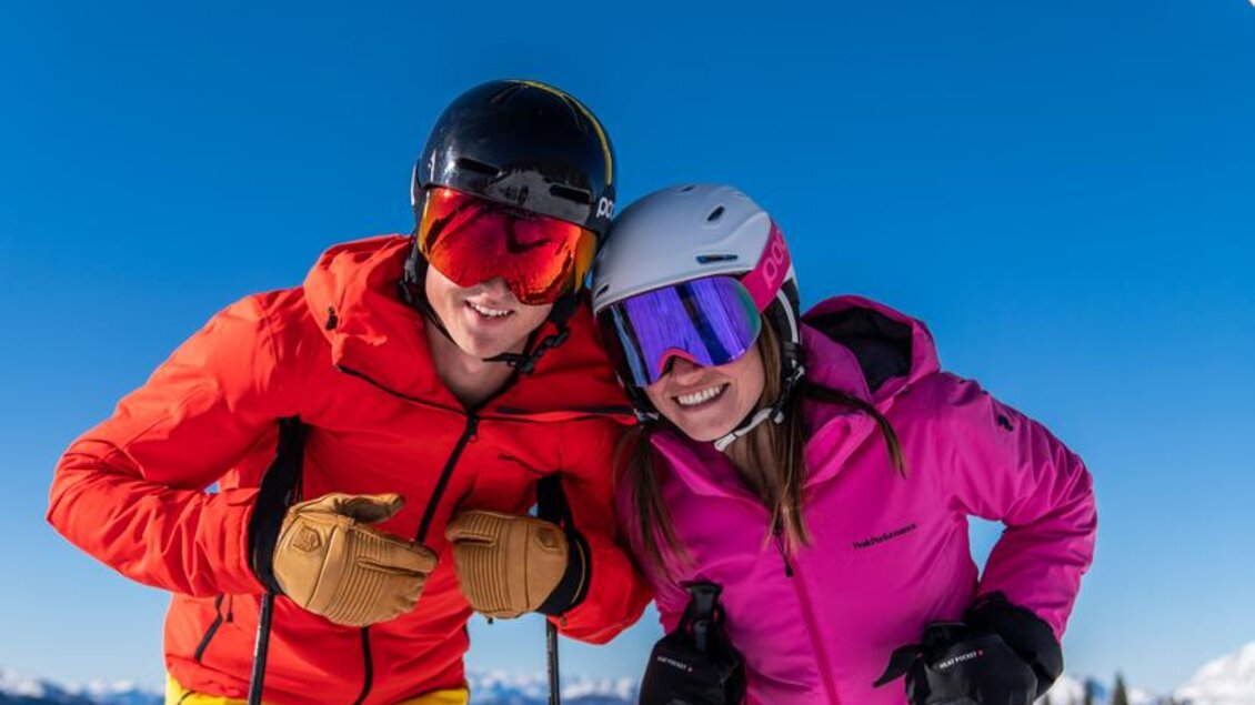 Zwei Skifahrer stehen fröhlich vor einem Schild mit der Aufschrift „FUNSLOPE“. Der Himmel ist klar und die Berge im Hintergrund sind schneebedeckt. | © Christine Höflehner