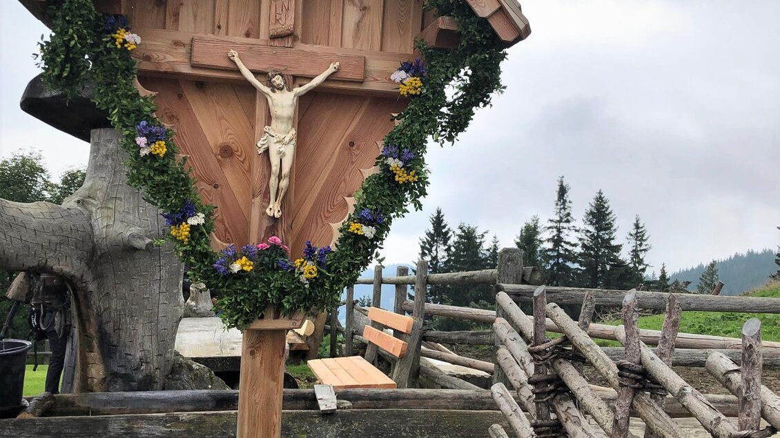 Ein hübscher Holzkreuz mit einem Christusbildnis, umgeben von Blumen. Im Hintergrund sind Bäume und ein Zaun zu sehen. | © Hofbauer Almhütte