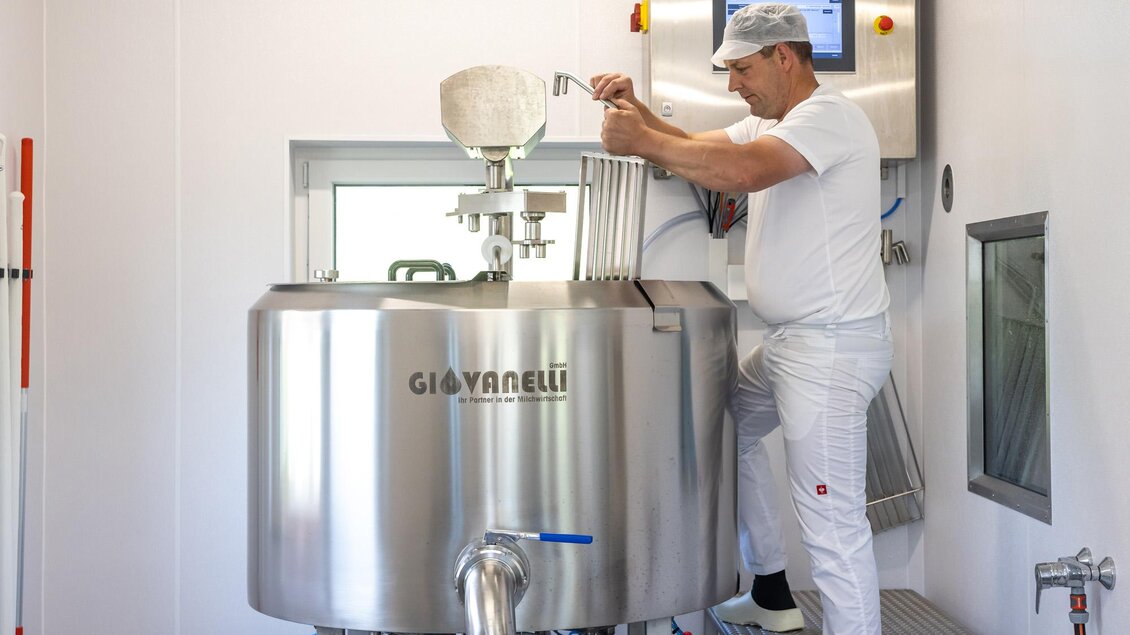 Ein Mann arbeitet an einer großen, silbernen Maschine in einer modernen Produktionsumgebung. Er fügt eine Zutat hinzu und beobachtet den Prozess aufmerksam. | © Martin Huber