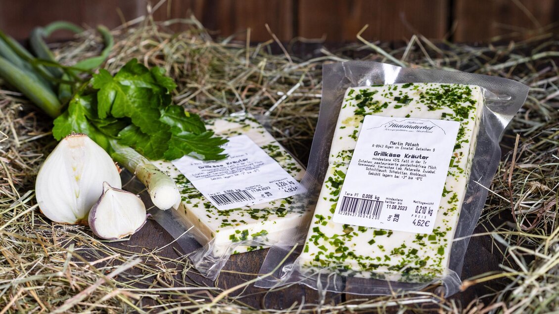 Zwei verpackte Käsestücke mit Kräutern liegen auf Heu. Daneben sind frische Zwiebeln und Sellerie zu sehen. | © Martin Huber