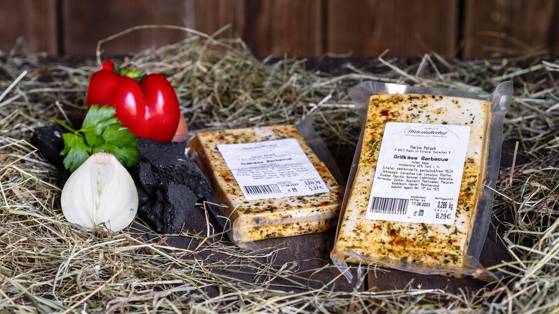 Zwei Käsepackungen liegen auf Heu, umgeben von frischen Gemüsezutaten wie Paprika und Zwiebeln. Der Hintergrund ist aus Holz, was eine rustikale Atmosphäre schafft. | © Martin Huber