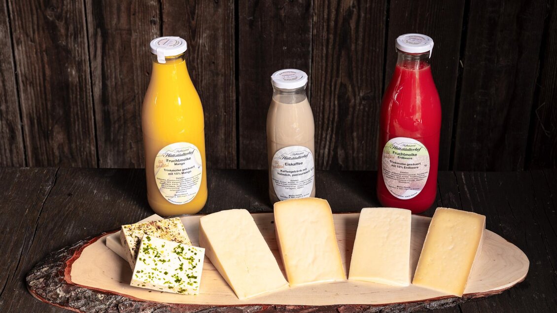Drei Flaschen Getränke stehen auf einem Holzteller, begleitet von verschiedenen Käsesorten. Der Hintergrund ist aus dunklem Holz, was eine rustikale Atmosphäre schafft. | © Martin Huber