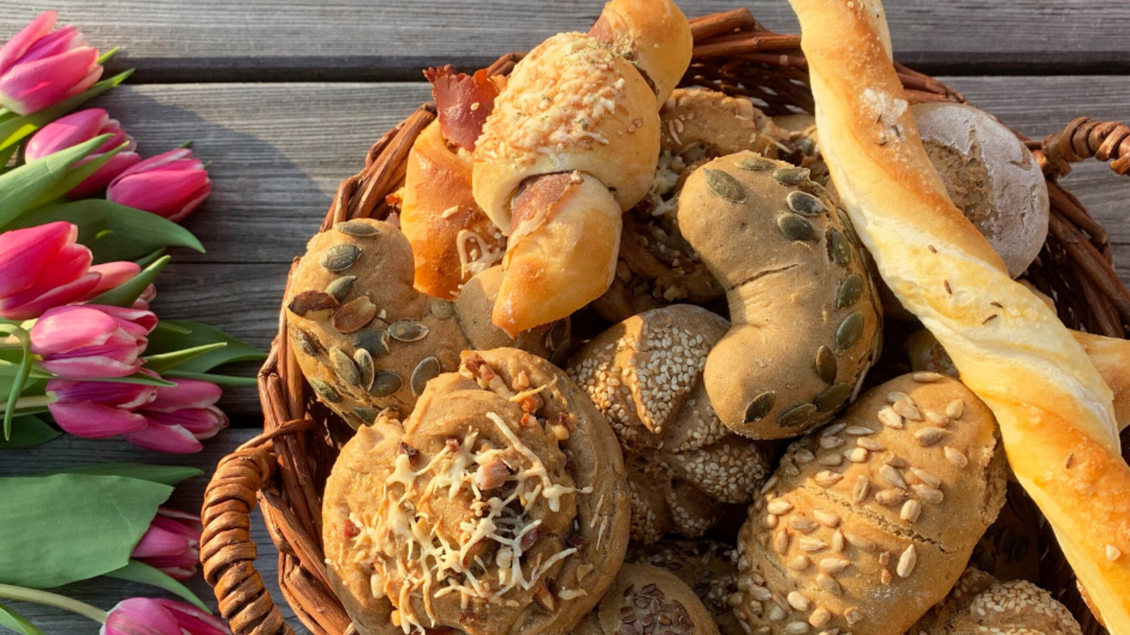 Ein Korb mit verschiedenen Brötchen und Gebäck liegt auf einem Holzuntergrund. Neben dem Korb liegen frische Tulpen. | © Bauernhof König - Familie König