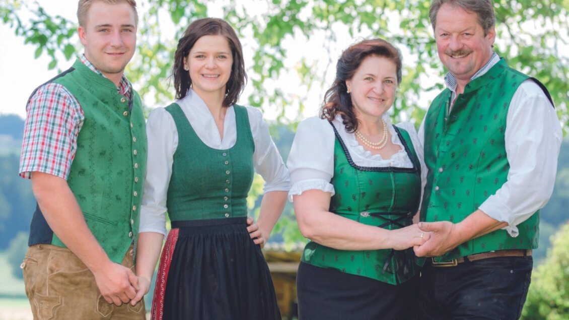Eine Familie in traditioneller Tracht steht lächelnd im Freien. Im Hintergrund sind grüne Blätter sichtbar, die eine fröhliche Atmosphäre schaffen. | © Bauernhof König - Familie König