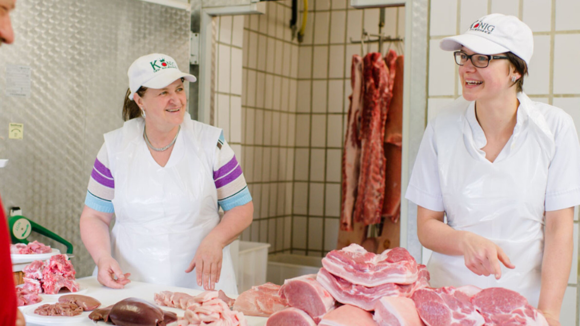 Zwei Frauen in einer Metzgerei lächeln und stehen hinter einem Tisch mit verschiedenen Fleischsorten. Im Hintergrund hängen Fleischstücke an Haken. | © Bauernhof König - Familie König