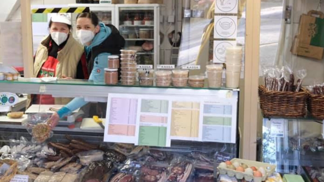 Zwei Verkäuferinnen stehen hinter einem Marktstand, der mit verschiedenen Lebensmitteln, Gläsern und Verpackungen gefüllt ist. Im Hintergrund sind Regale mit weiteren Produkten sichtbar. | © Bauernhof König - Familie König