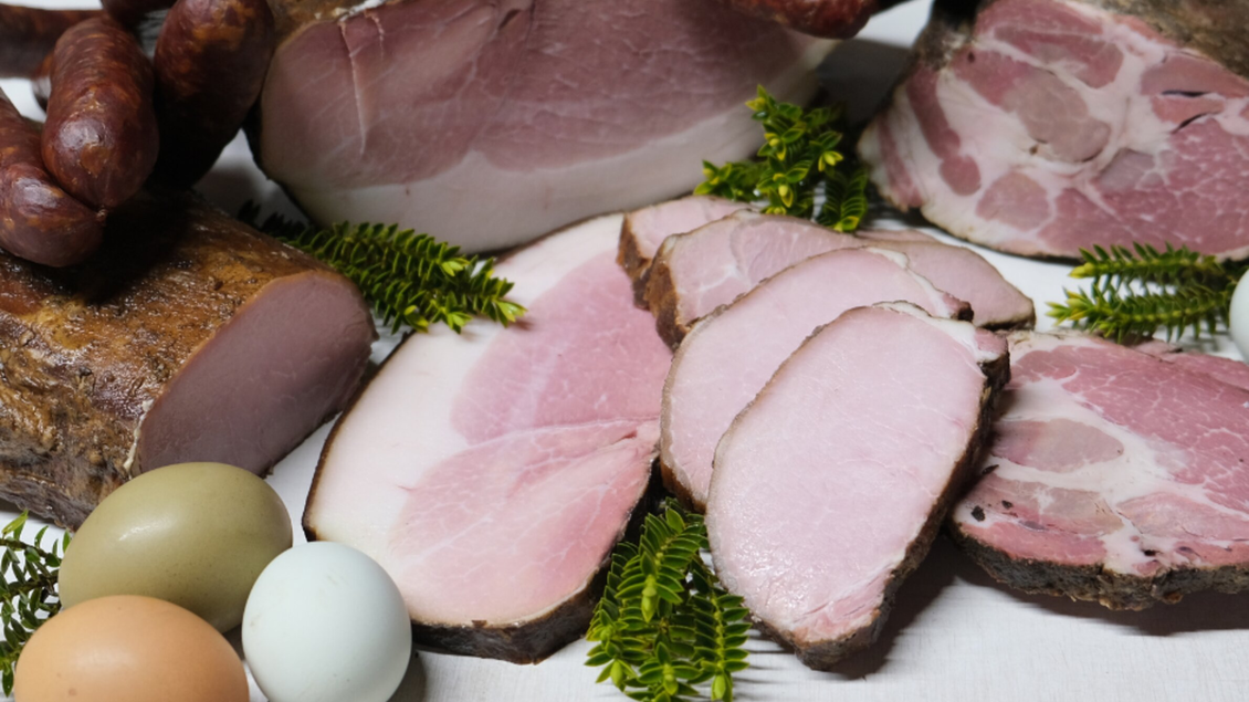 Eine Auswahl an Fleischwaren, einschließlich geschnittenem Schinken und Wurst, umgeben von grünen Farnen und Eiern. Es zeigt frische, handwerklich hergestellte Delikatessen. | © Bauernhof König - Familie König