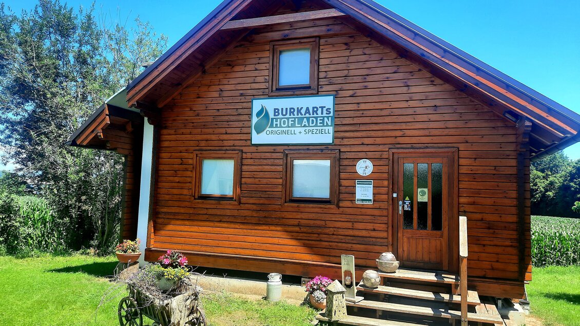 Ein rustikales Holzhaus mit einem Schild, das "Burkarts Hofladen" anzeigt. Vor dem Eingang stehen Blumenkübel und eine alte Wagenrad-Deko. | © Sigrid Burkart