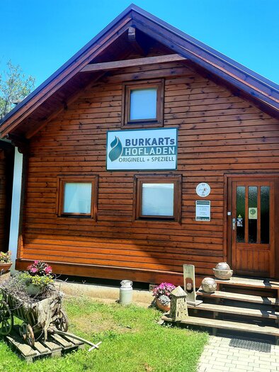 Ein rustikales Holzhaus mit einem Schild, das "Burkarts Hofladen" anzeigt. Vor dem Eingang stehen Blumenkübel und eine alte Wagenrad-Deko. | © Sigrid Burkart