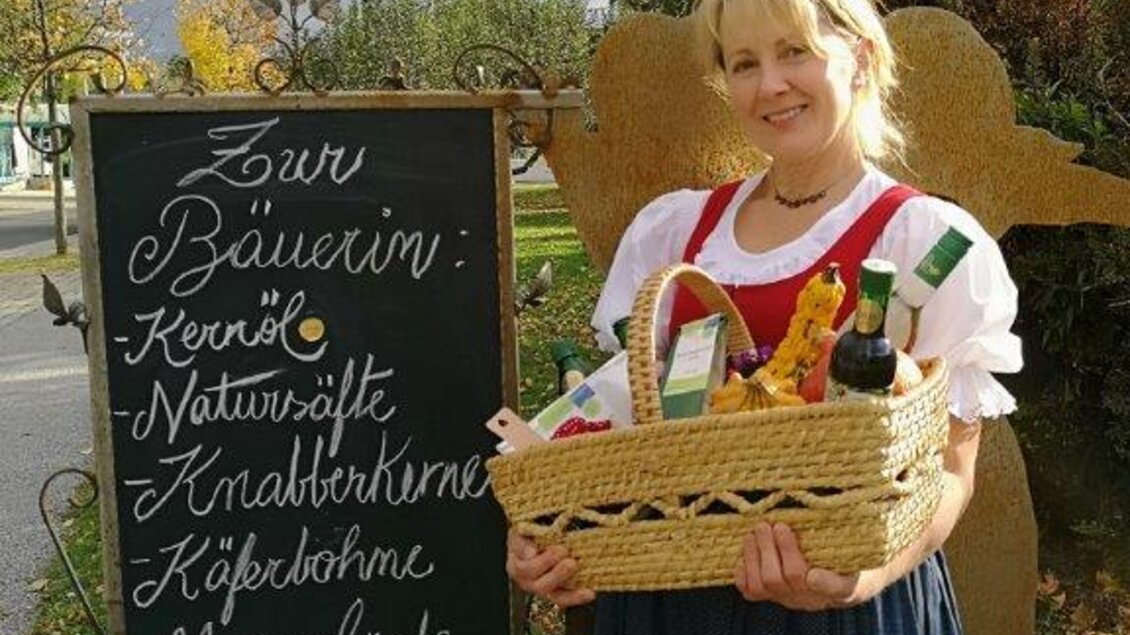 Eine Frau in traditioneller Tracht hält einen Korb mit regionalen Produkten. Im Hintergrund steht ein Schild mit Angeboten von einer Bäuerin. | © Familie Brugner