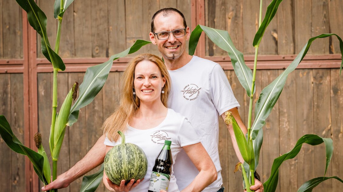 Ein Paar steht zwischen hohen Pflanzen und hält eine Melone sowie eine Flasche in der Hand. Der Hintergrund zeigt eine Holzwand, die eine ländliche Atmosphäre vermittelt. | © Region Graz - Mias Photoart